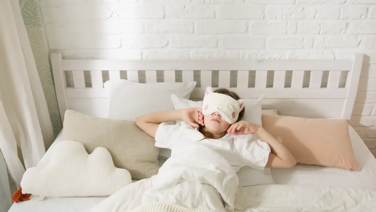 A young girl resting in bed with a cute sleep mask surrounded by cozy pillows.