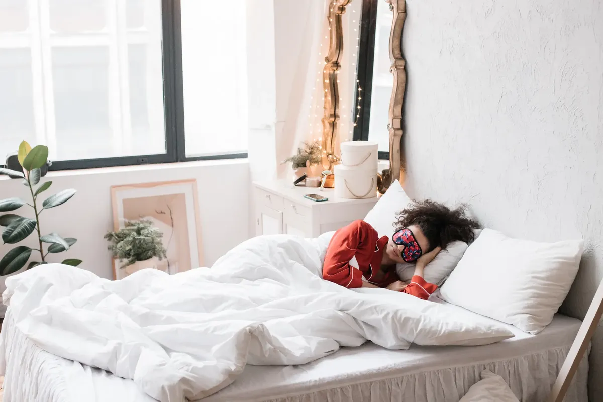 睡眠マスクをかけた女性が、柔らかい白いベッドに横たわり、明るい部屋に室内の植物が囲んでいます。