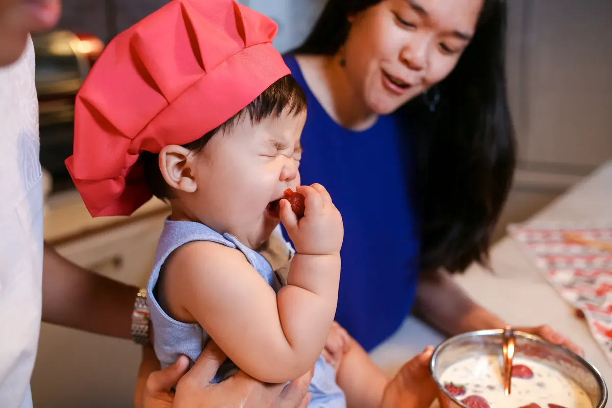 Baby in chef hat enjoys strawberries with family in kitchen. Perfect for depicting family cooking fun.
