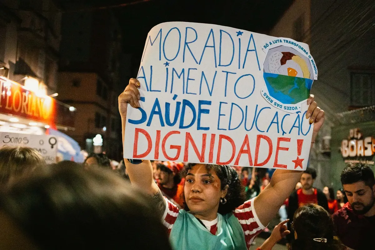 Woman holding sign demanding housing, food, health, education, and dignity at a protest.